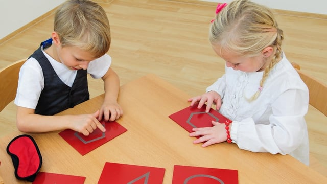 zwei Kinder sitzen am Tisch und fahren mit den Fingern Formen auf Tastplatten nach